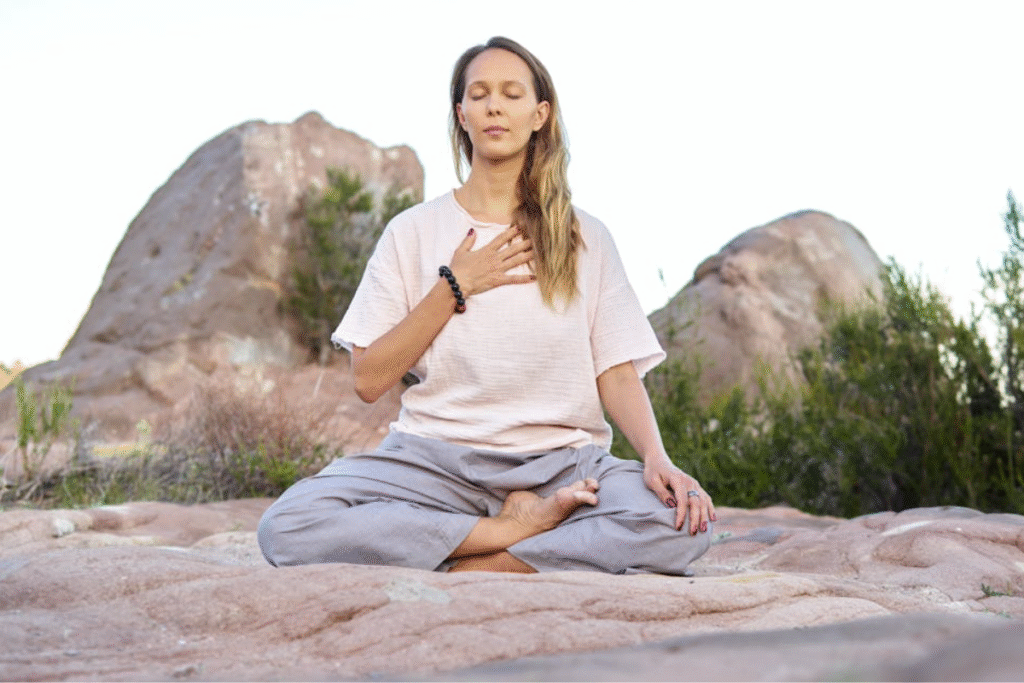 A woman meditating peacefully outdoors in a natural, rocky environment, with one hand on her heart.