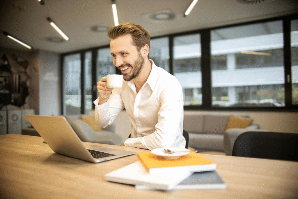 A happy, smiling man in a white shirt enjoying a cup of coffee while working on his laptop in a modern office.