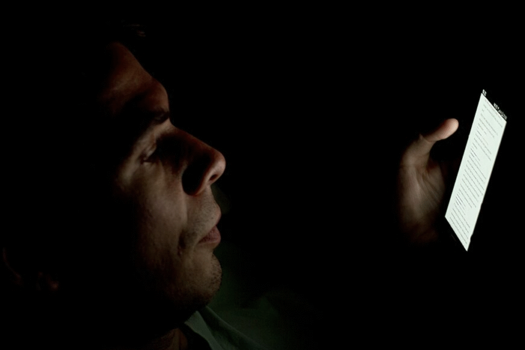 A man's face in a dark room, illuminated solely by the bright white light from the smartphone he is holding.